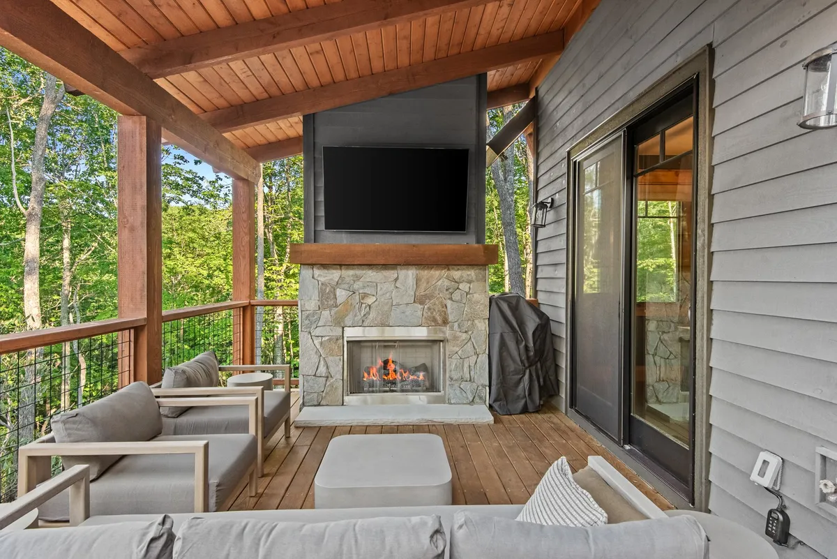 A cozy outdoor living space featuring a stone fireplace, comfortable seating, and a flat-screen TV, all nestled on a deck overlooking surrounding trees. The inviting setting suggests evenings spent together outdoors, enjoying movies or sharing stories by the fire.