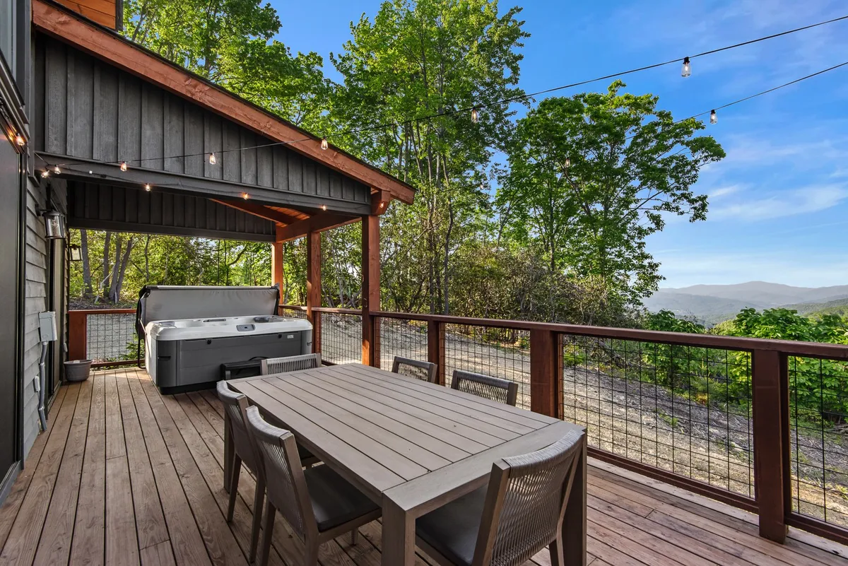 An inviting outdoor space featuring a hot tub, dining table, and serene mountain views, all surrounded by trees. The mood is relaxed and offers a perfect setting for unwinding after a day of exploration.