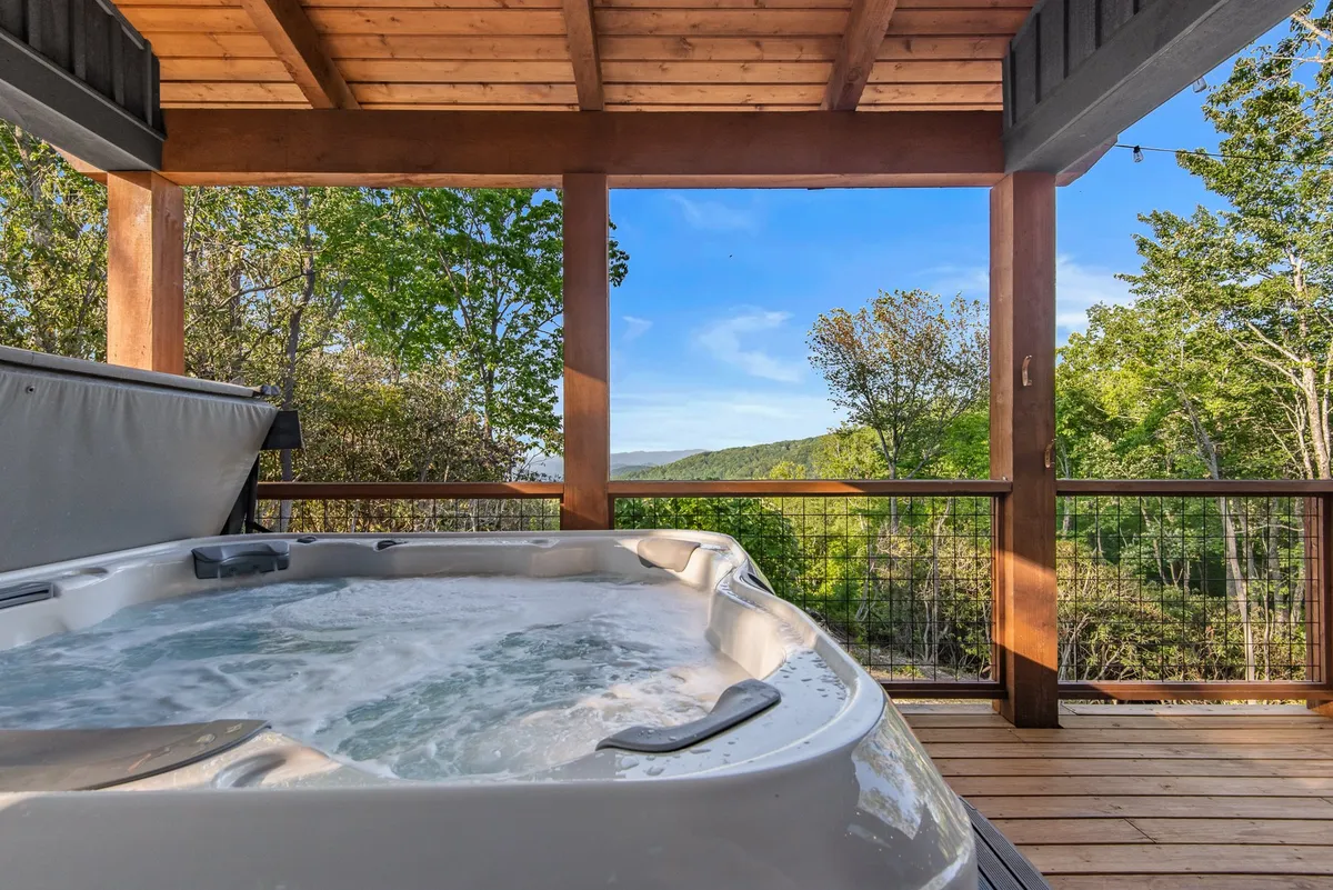 A serene exterior shot featuring a spacious wooden deck with a six-person hot tub overlooking expansive mountain scenery. The setting exudes tranquility and invites guests to unwind, with hints of the sunset and the promise of restorative soaks after mountain adventures.