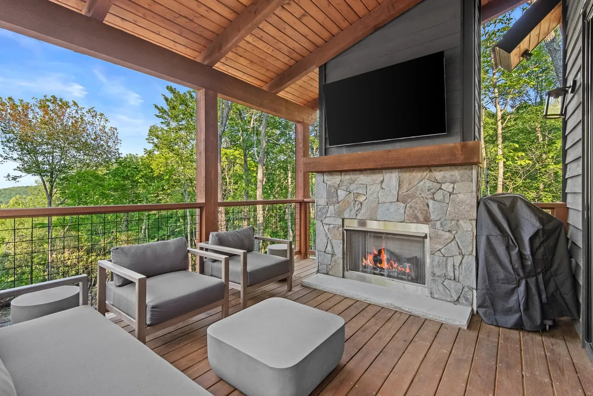 A partially covered outdoor deck with a welcoming seating area surrounding a modern gas fireplace and an outdoor television. The space is accentuated by surrounding greenery and a sense of privacy, inviting guests to enjoy evenings outdoors, be it for movie nights or relaxed gatherings under the stars.