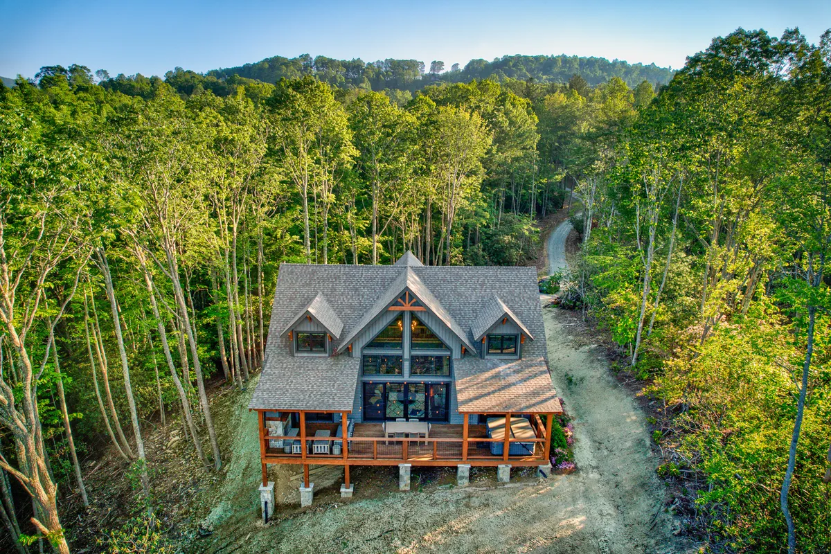 This image showcases a charming log cabin with a spacious front porch surrounded by trees. The driveway leads into the property, emphasizing a welcoming and natural setting perfect for outdoor activities and walks.
