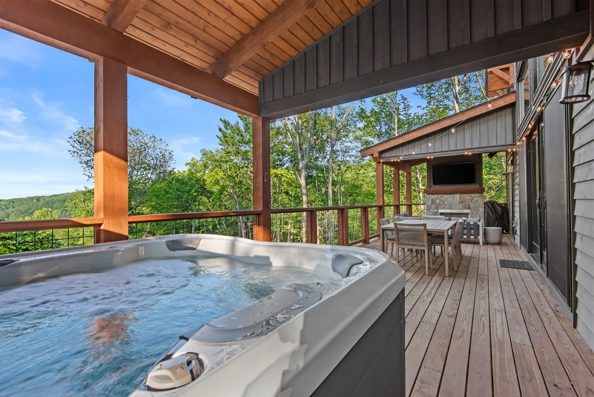 This exterior scene showcases a private deck featuring a luxurious hot tub, an outdoor dining area, and sturdy wooden beams. The setting is surrounded by natural beauty, offering sweeping views and a tranquil atmosphere perfect for relaxation or socializing. The hot tub is centrally featured, highlighting its capacity for group comfort while the outdoor seating emphasizes the home’s connection to the surrounding mountains.