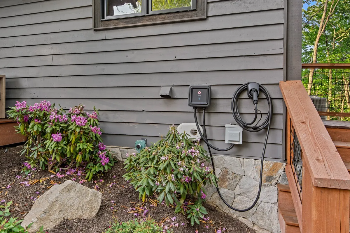 This exterior image features a welcoming porch surrounded by purple flowering shrubs and an EV charging station. Wooden steps invite you up, and the setting is lush with greenery, indicating both environmental consciousness and a connection to the natural landscape.