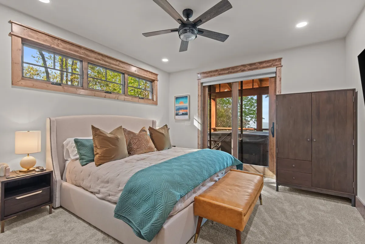 A serene master bedroom with a large, comfortable bed adorned with decorative pillows. A nightstand with a lamp sits beside the bed, and glass doors open to a scenic view outside, letting in plenty of natural light for a peaceful atmosphere.