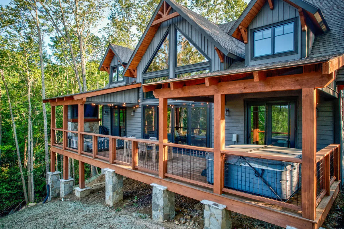 A spacious wooden deck surrounded by lush trees, featuring a hot tub and large windows looking into the interior. The deck offers ample room for outdoor relaxation and play, making it ideal for both people and pets. The natural setting is inviting for families with dogs to enjoy fresh mountain air and freedom.