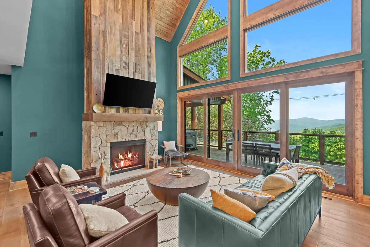A welcoming open-concept living room featuring plush, comfortable sofas arranged around a striking indoor fireplace. Large windows allow natural light to flood the space and frame impressive mountain views, while the arrangement encourages conversation and relaxation. The scene suggests warmth, togetherness, and relaxation, ideal for both cozy evenings and lively gatherings.