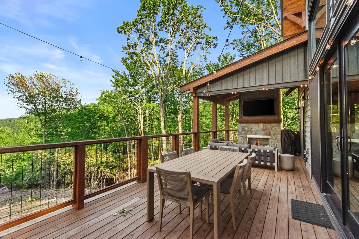 The image presents a spacious outdoor area with a large dining table, stone fireplace, comfortable seating, and mature trees surrounding the space. The area is designed for gatherings, outdoor meals, and cozy evenings spent by the fire, all while immersed in nature.